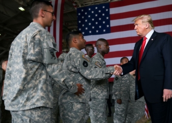 El expresidente Donald Trump saluda a un grupo de militares uniformados frente a una gran bandera de los Estados Unidos en un hangar oficial.