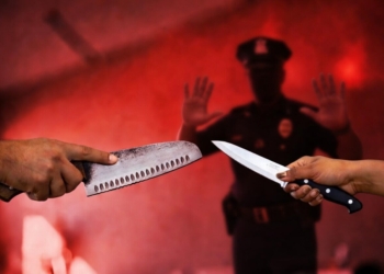 Dos personas enfrentándose con cuchillos mientras un policía intenta intervenir al fondo en una escena de tensión y violencia.