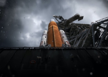 Cohete espacial en plataforma de lanzamiento bajo cielo nublado y lluvioso, con estructura de torre al fondo.