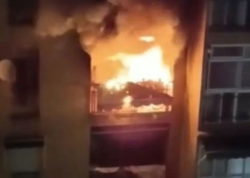 Incendio en el interior de un edificio visto desde una ventana, con llamas intensas y humo en un entorno urbano.