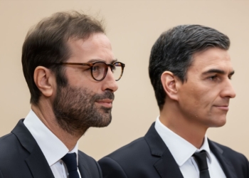 Carlos Cuerpo y Pedro Sánchez con traje en perfil con expresión seria en un entorno neutro