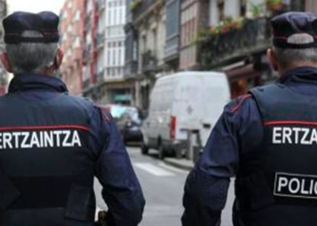 Dos agentes de la Ertzaintza patrullando de espaldas por una calle urbana con vehículos y edificios alrededor.