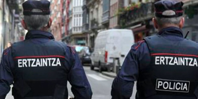 Dos agentes de la Ertzaintza patrullando de espaldas por una calle urbana con vehículos y edificios alrededor.
