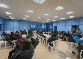 Sala de espera con numerosas personas sentadas en sillas de plástico en un espacio interior tipo oficina o centro administrativo.