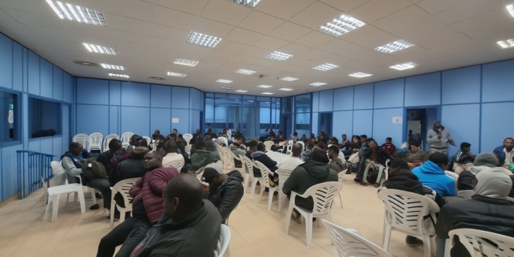 Sala de espera con numerosas personas sentadas en sillas de plástico en un espacio interior tipo oficina o centro administrativo.
