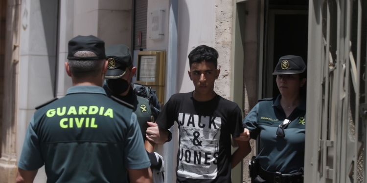 Agentes de la Guardia Civil trasladando a un joven detenido a la salida de un edificio en un entorno urbano.