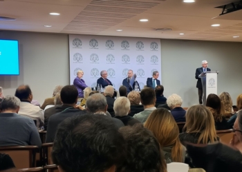 Conferencia en sala con público asistente y varios ponentes en una mesa, mientras un orador interviene desde un atril.