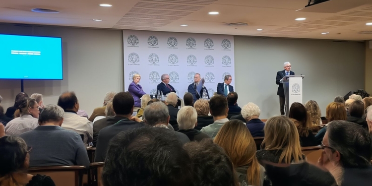 Conferencia en sala con público asistente y varios ponentes en una mesa, mientras un orador interviene desde un atril.