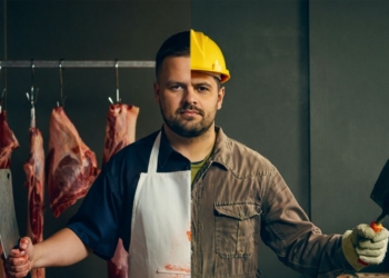 Montaje irónico sobre el pluriempleo con Imagen dividida de un hombre mitad carnicero con piezas de carne colgadas y mitad obrero con casco, sosteniendo herramientas en ambos lados.