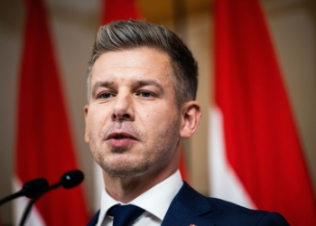 Primer plano de Peter Magyar hablando en rueda de prensa con banderas rojas desenfocadas al fondo.