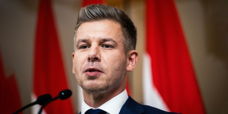 Primer plano de Peter Magyar hablando en rueda de prensa con banderas rojas desenfocadas al fondo.