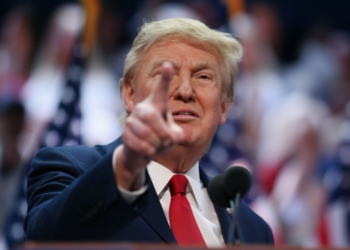 Donald Trump señalando con el dedo durante un discurso, con banderas de fondo desenfocadas.