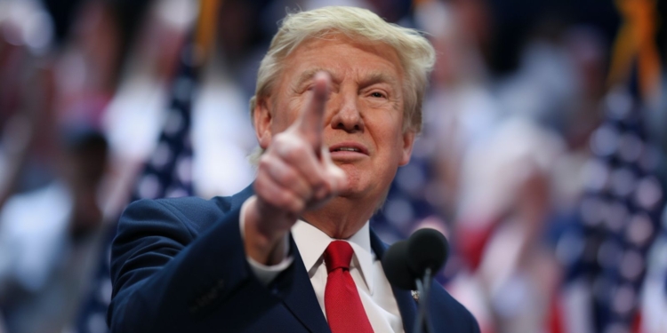 Donald Trump señalando con el dedo durante un discurso, con banderas de fondo desenfocadas.