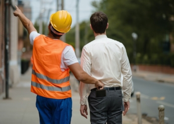 Un trabajador con casco señala hacia adelante mientras roba la cartera a un hombre que camina a su lado por la calle.