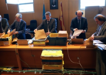 Varios funcionarios revisan y clasifican sobres y documentos en una mesa durante un proceso de recuento electoral en una sala oficial.