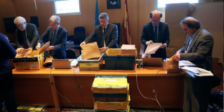 Varios funcionarios revisan y clasifican sobres y documentos en una mesa durante un proceso de recuento electoral en una sala oficial.
