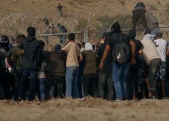 Grupo de personas intentando cruzar una valla con alambre de espino en una zona árida, mientras otras ya están trepando o pasando al otro lado.