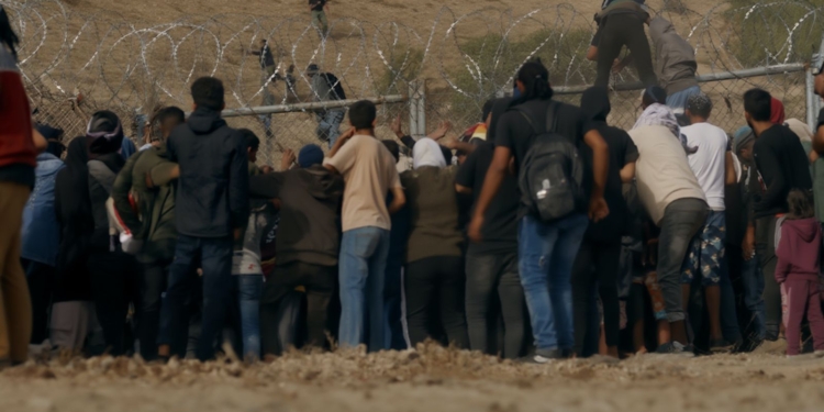 Grupo de personas intentando cruzar una valla con alambre de espino en una zona árida, mientras otras ya están trepando o pasando al otro lado.