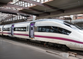 Tren de alta velocidad blanco con una franja morada detenido en un andén de estación, con puertas abiertas y estructura metálica cubierta sobre las vías.