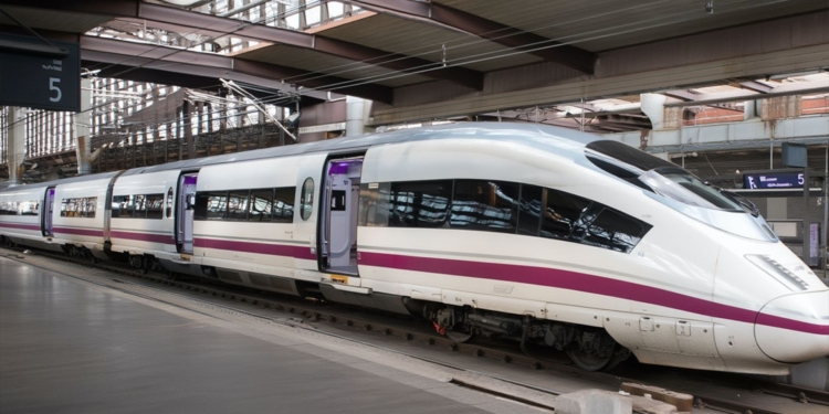 Tren de alta velocidad blanco con una franja morada detenido en un andén de estación, con puertas abiertas y estructura metálica cubierta sobre las vías.