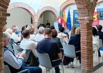 Evento de Startup Olé en Marbella, en un patio interior con arcos de ladrillo, donde varias personas están sentadas escuchando a una ponente frente a banderas y una pantalla de presentación.