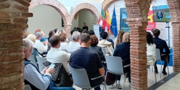 Evento de Startup Olé en Marbella, en un patio interior con arcos de ladrillo, donde varias personas están sentadas escuchando a una ponente frente a banderas y una pantalla de presentación.