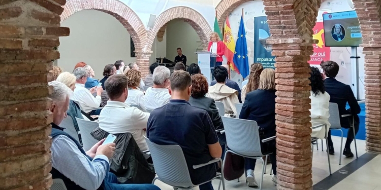 Evento de Startup Olé en Marbella, en un patio interior con arcos de ladrillo, donde varias personas están sentadas escuchando a una ponente frente a banderas y una pantalla de presentación.