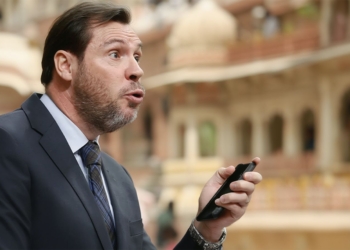 El Ministro Óscar Puente hablando mientras sostiene un teléfono móvil, con gesto expresivo, en un entorno arquitectónico con arcos desenfocados al fondo.