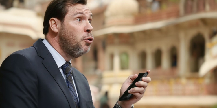 El Ministro Óscar Puente hablando mientras sostiene un teléfono móvil, con gesto expresivo, en un entorno arquitectónico con arcos desenfocados al fondo.