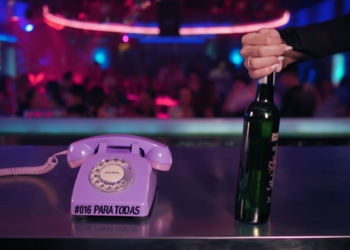 Teléfono fijo de estilo retro con el texto “#016 PARA TODAS” sobre una barra, mientras una mano sostiene una botella en un ambiente de discoteca con luces de colores y gente desenfocada al fondo.