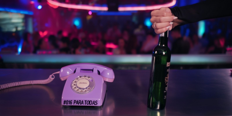 Teléfono fijo de estilo retro con el texto “#016 PARA TODAS” sobre una barra, mientras una mano sostiene una botella en un ambiente de discoteca con luces de colores y gente desenfocada al fondo.