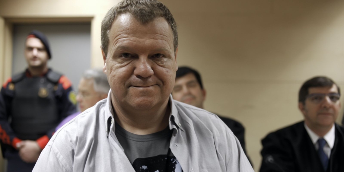 Joan Vila Dilmé conocido como el Asesino de Olot, en primer plano dentro de una sala judicial, con varias personas sentadas detrás y un agente uniformado al fondo.