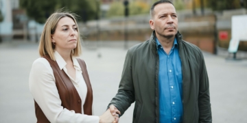 María Guardiola y Óscar Fernández de pie en la calle, dándose la mano con gesto serio mientras miran hacia el frente.