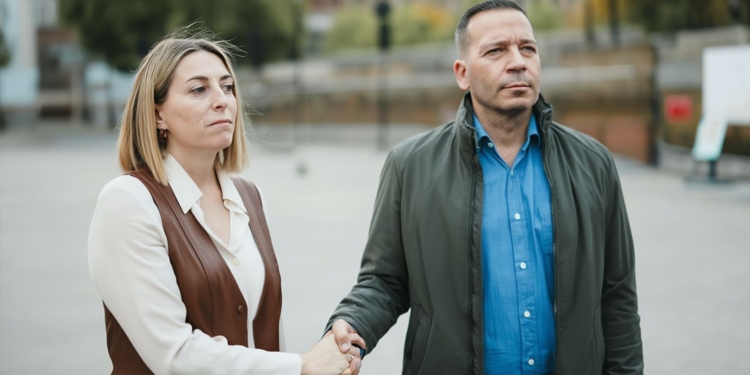 María Guardiola y Óscar Fernández de pie en la calle, dándose la mano con gesto serio mientras miran hacia el frente.