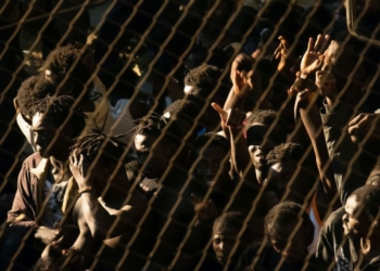 Un grupo numeroso de personas detrás de una valla metálica levanta las manos y se agolpa, en un ambiente tenso y con iluminación dura.