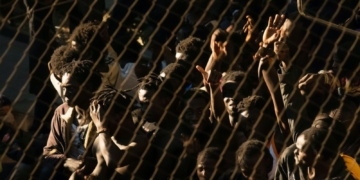 Un grupo numeroso de personas detrás de una valla metálica levanta las manos y se agolpa, en un ambiente tenso y con iluminación dura.