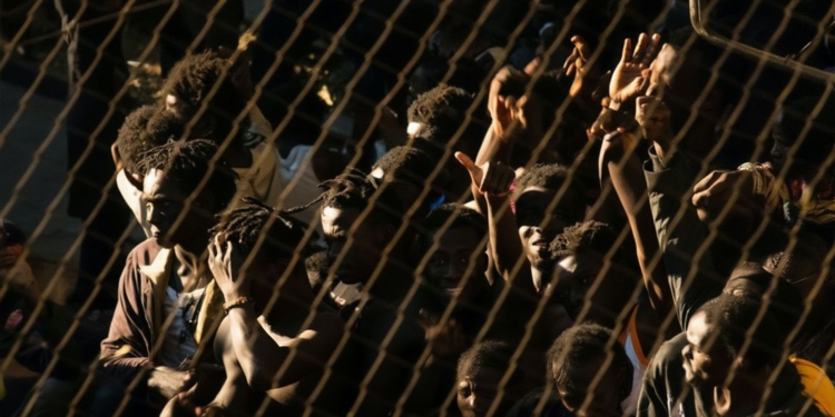 Un grupo numeroso de personas detrás de una valla metálica levanta las manos y se agolpa, en un ambiente tenso y con iluminación dura.