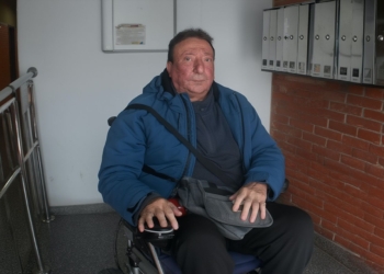 Pepe en silla de ruedas dentro de un portal, junto a buzones de correo, mira al frente con expresión seria.