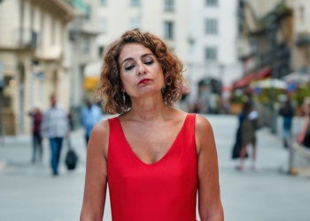 Maria Jesús Montero con vestido rojo de tirantes, con expresión seria y los ojos cerrados, de pie en una calle urbana con gente desenfocada al fondo.