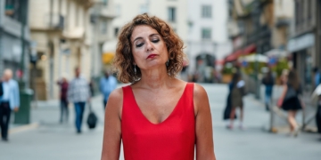 Maria Jesús Montero con vestido rojo de tirantes, con expresión seria y los ojos cerrados, de pie en una calle urbana con gente desenfocada al fondo.