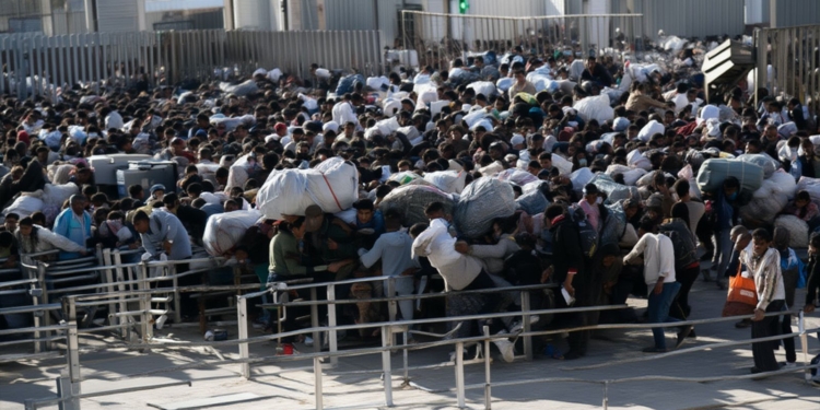 Gran multitud de personas concentradas en un paso fronterizo o acceso controlado, muchas cargando bolsas y pertenencias voluminosas, avanzando de forma aglomerada entre vallas metálicas.