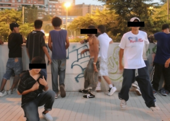 Grupo de jóvenes en un entorno urbano, algunos apoyados en un muro con grafitis; varios rostros están cubiertos con franjas negras para ocultar la identidad.