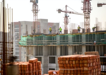 Obreros con chalecos de seguridad trabajan en la parte superior de un edificio en construcción, rodeados de grúas y estructuras de hormigón, con pilas de ladrillos en primer plano y edificios urbanos al fondo.