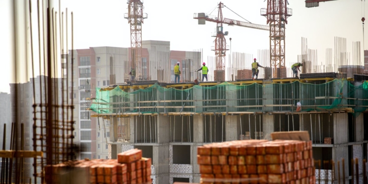 Obreros con chalecos de seguridad trabajan en la parte superior de un edificio en construcción, rodeados de grúas y estructuras de hormigón, con pilas de ladrillos en primer plano y edificios urbanos al fondo.