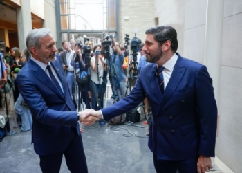 Alejandro Nolasco y Jorge Azcón con traje se dan la mano en un edificio mientras periodistas y cámaras observan al fondo.