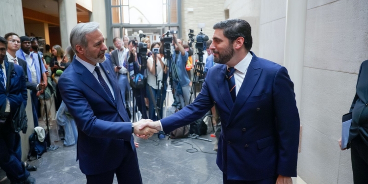 Alejandro Nolasco y Jorge Azcón con traje se dan la mano en un edificio mientras periodistas y cámaras observan al fondo.