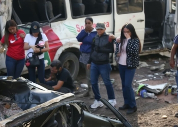 Personas observan los restos de un vehículo tras un accidente, con un autobús dañado al fondo en Colombia.