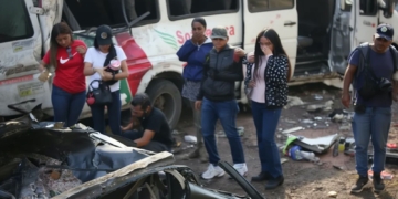 Personas observan los restos de un vehículo tras un accidente, con un autobús dañado al fondo en Colombia.