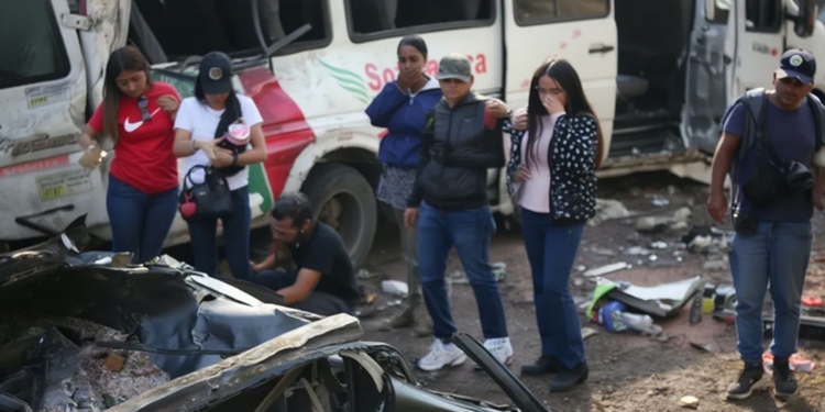 Personas observan los restos de un vehículo tras un accidente, con un autobús dañado al fondo en Colombia.
