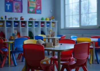 Aula infantil vacía con mesas y sillas de colores y estanterías con juguetes al fondo.